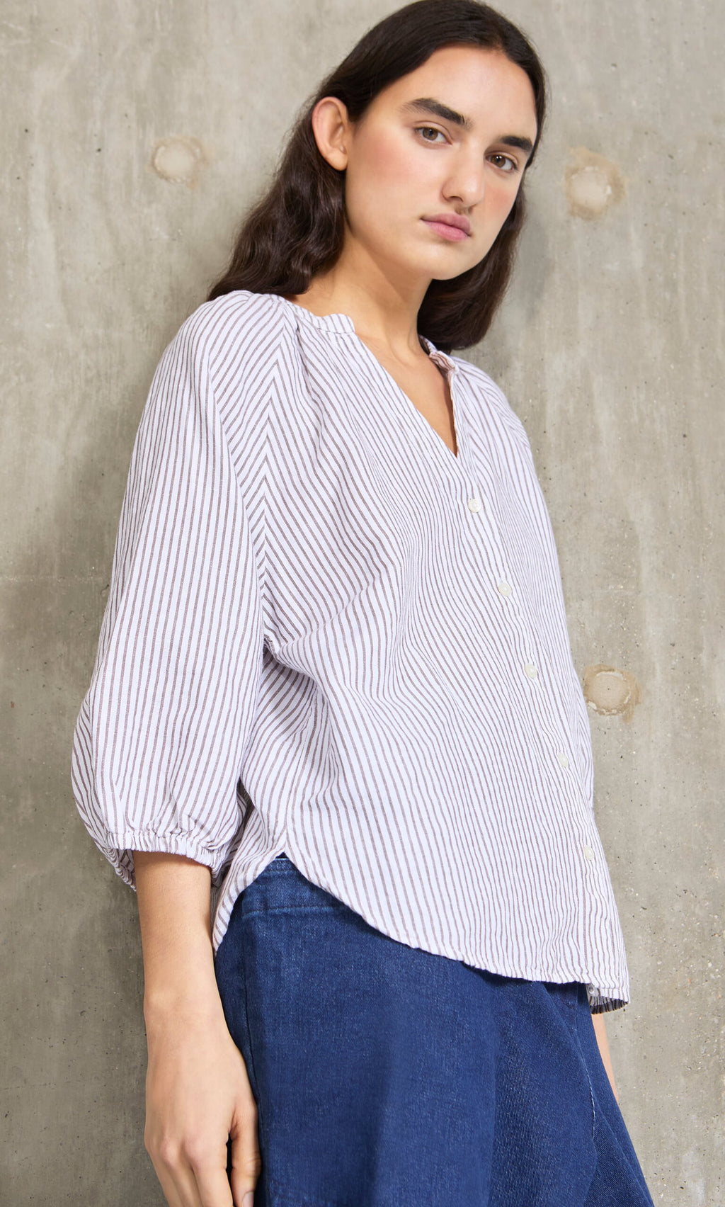 Plümo- Person in a relaxed fit Brisley Blouse and blue pants stands against a textured concrete wall, looking at the camera.