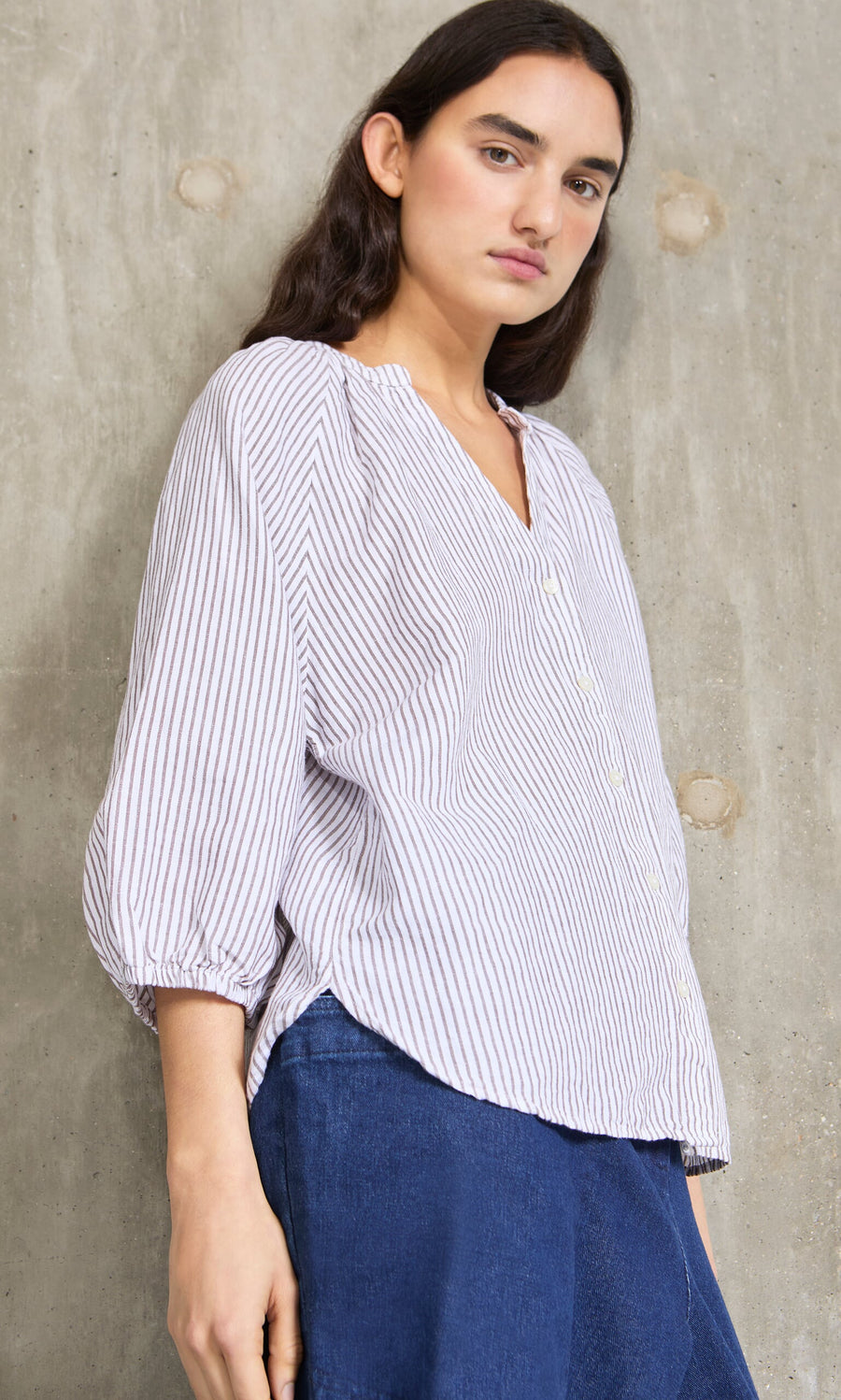 Plümo- Person in a relaxed fit Brisley Blouse and blue pants stands against a textured concrete wall, looking at the camera.