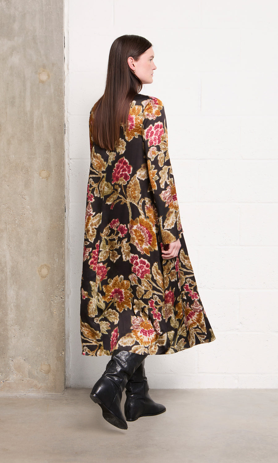 Plümo  Woman in the Brunn Dress and black boots stands sideways near a concrete and white brick wall.