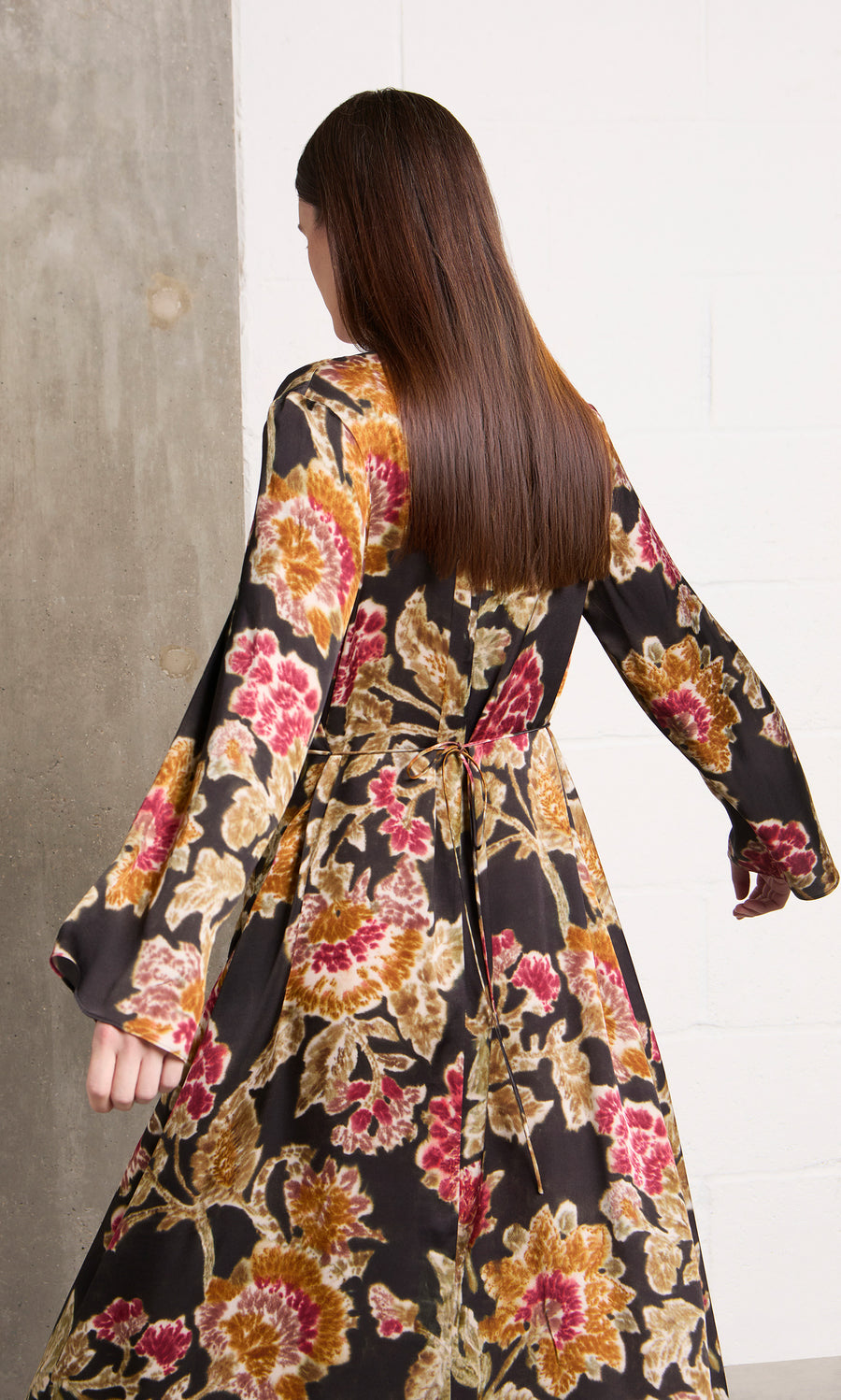 Plümo  Woman with long brown hair in the Brunn Dress, walking by a concrete and white wall.