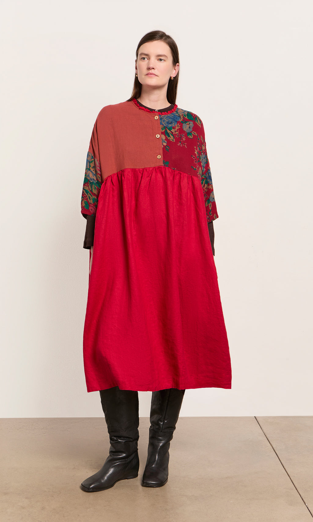 Plümo  Woman in the Burns Patchwork Dress, midi-length with florals, styled with black boots against a plain backdrop.