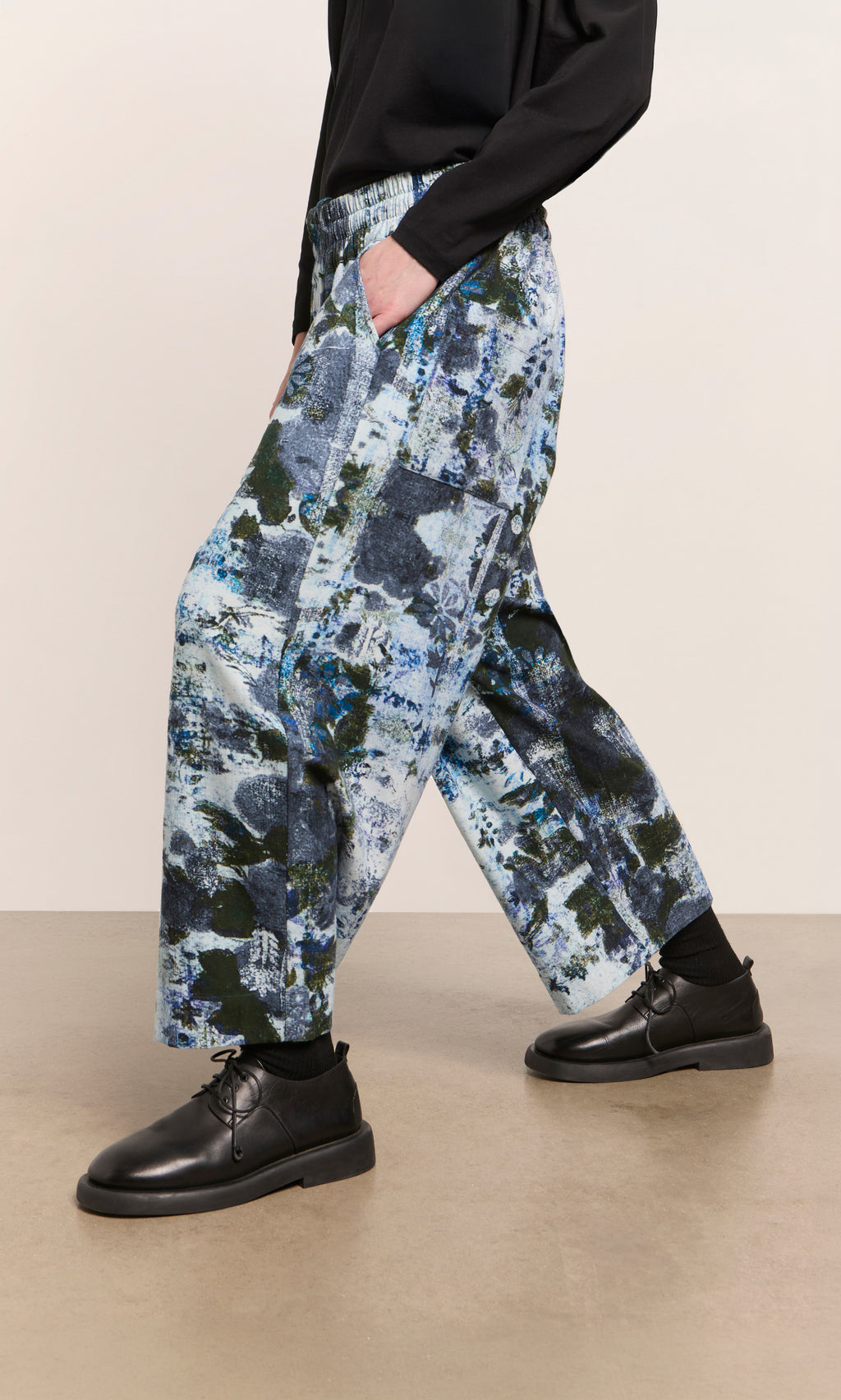 Plümo  Person in Caccia Pants with a wintery print, black top and shoes, standing on a light brown floor.