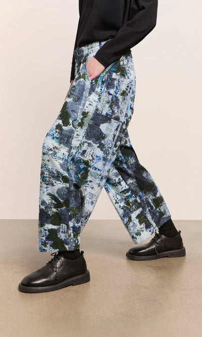 Plümo  Person in Caccia Pants with a wintery print, black top and shoes, standing on a light brown floor.