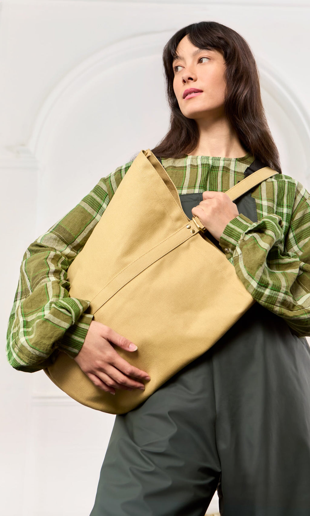 Plümo- Woman in a green plaid top and grey overalls carries a weatherproof canvas tool bag, looking to the side.