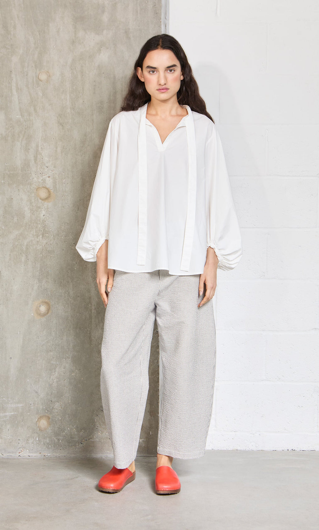Plümo- Woman stands indoors by a concrete wall in a loose white blouse and high waist Passanti Pants, paired with red shoes.