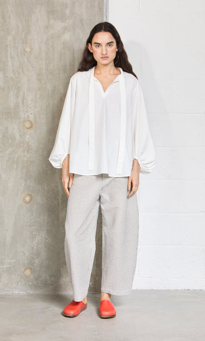 Plümo- Woman stands indoors by a concrete wall in a loose white blouse and high waist Passanti Pants, paired with red shoes.