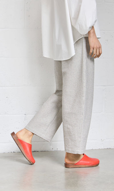 Plümo- Person in loose gray pants, white shirt, and Carlie Clogs stands against a white wall on a concrete floor.