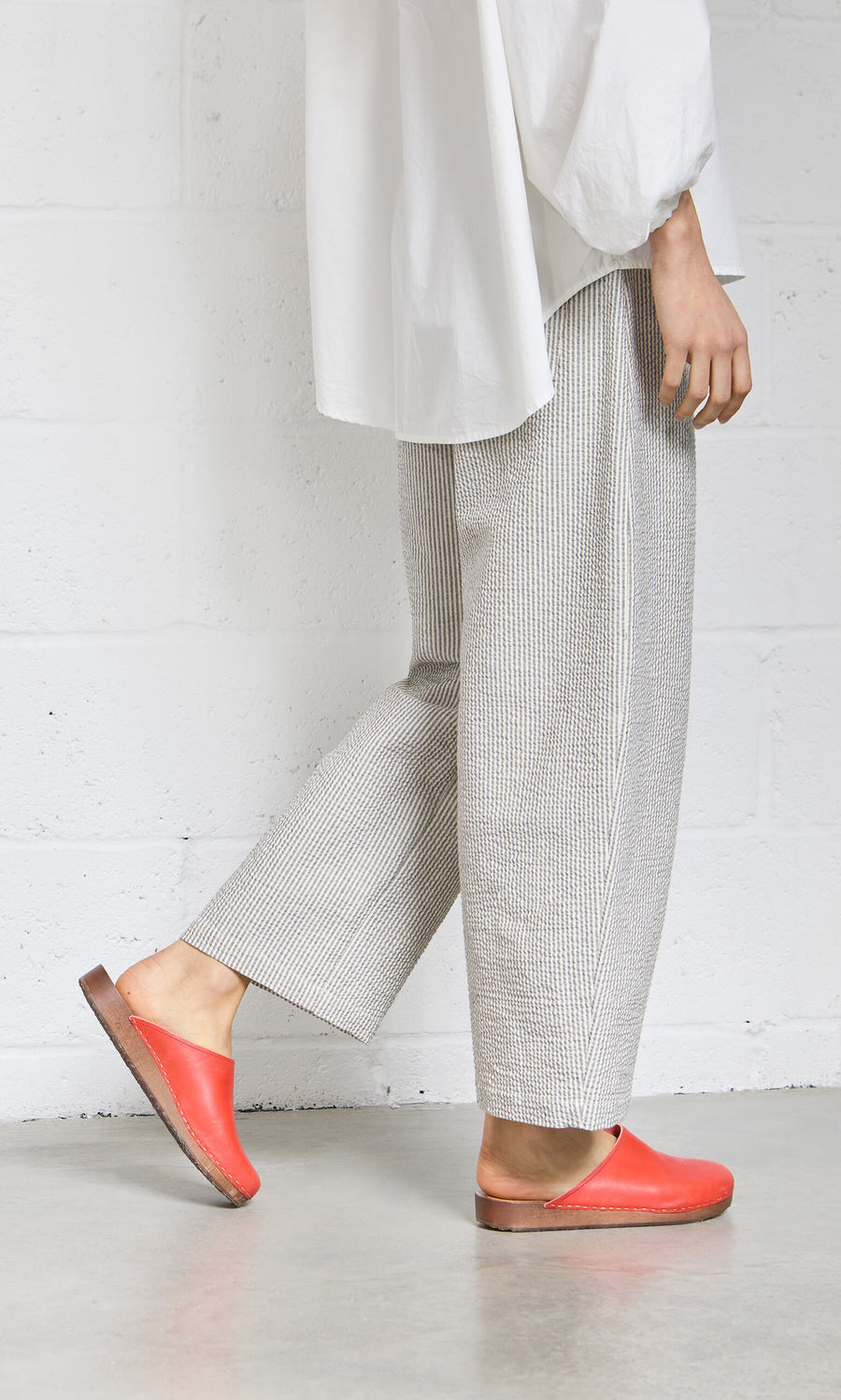 Plümo- Person in loose gray pants, white shirt, and Carlie Clogs stands against a white wall on a concrete floor.
