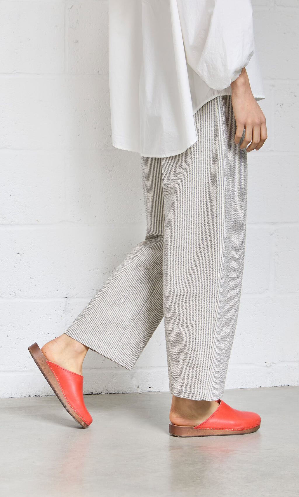 Plümo- A person in Passanti Pants, a white shirt, and red slip-on shoes stands against a white wall.