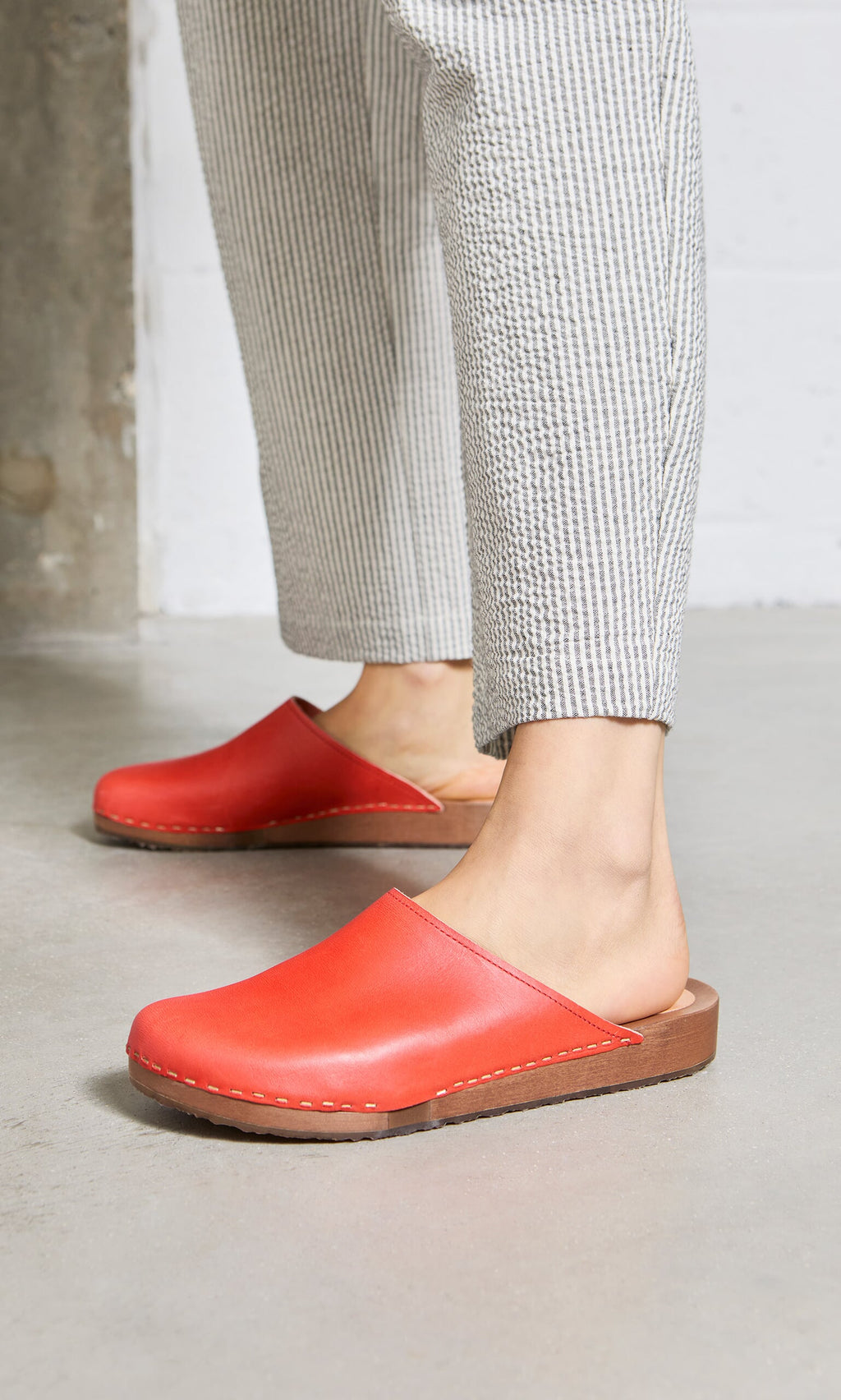 Plümo- Person wearing Carlie Clogs in red vegetable-tanned leather with light gray striped pants on a concrete floor.