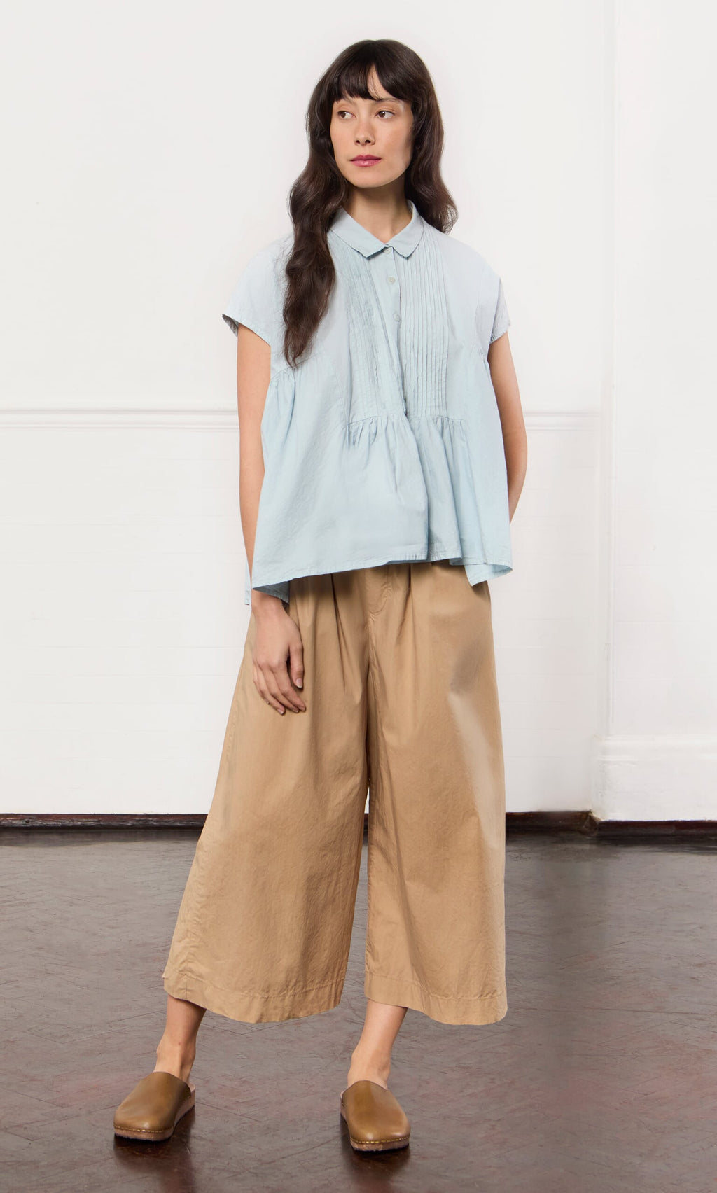 Plümo- Woman wears Howland Pants—sustainably made, wide-leg cotton pull-ons—with a light blue blouse against a white wall.