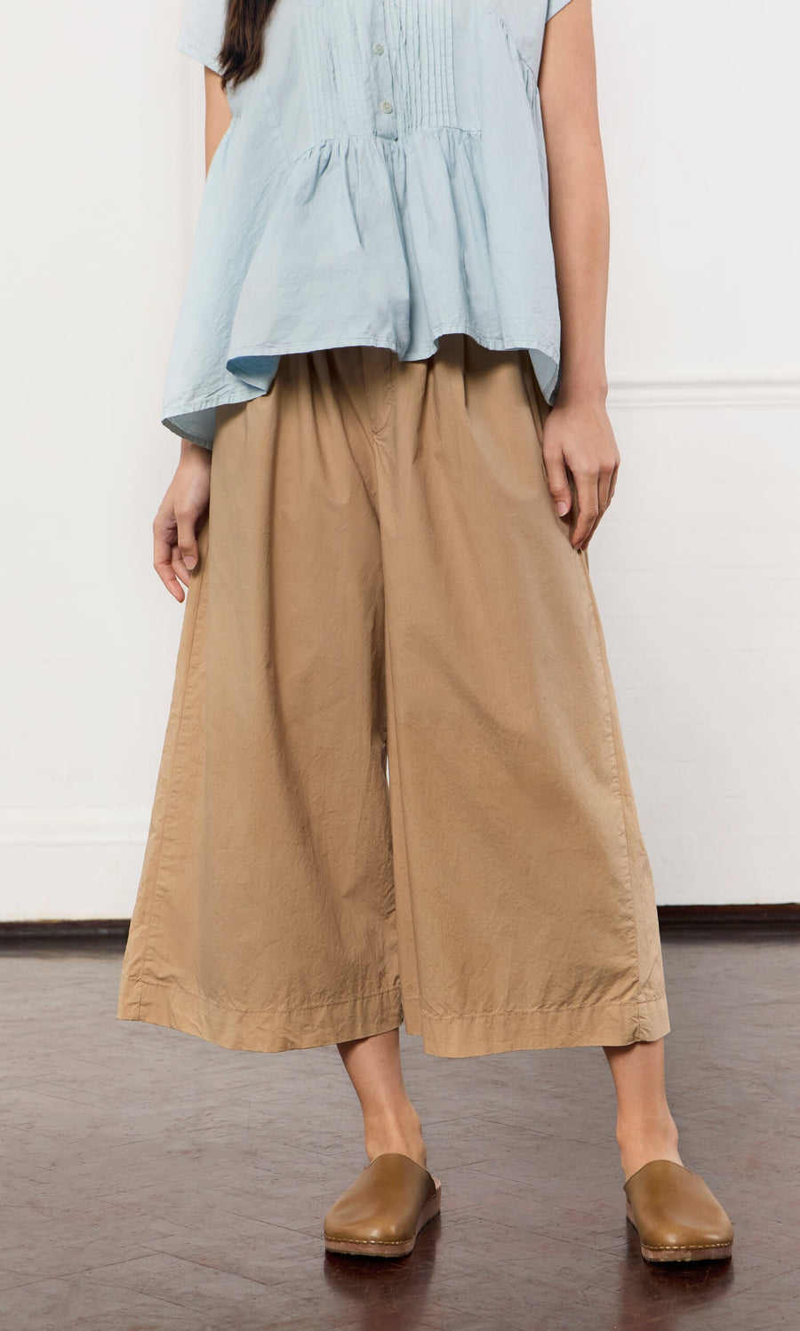 Plümo- Woman wearing a light blue blouse, Howland Pants in wide-leg cotton, and brown clogs standing indoors on a dark floor.