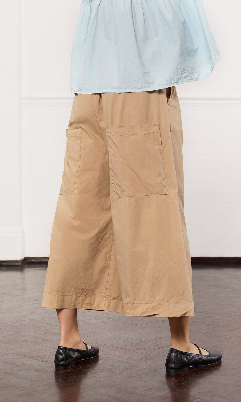 Plümo- Person wearing Howland Pants in tan, wide-leg silhouette, with black flats and a light blue top, standing indoors.