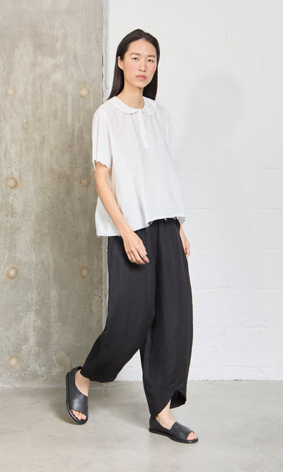Plümo- Someone in a loose white top and Cazalla Pants - Black stands against a concrete and white wall.
