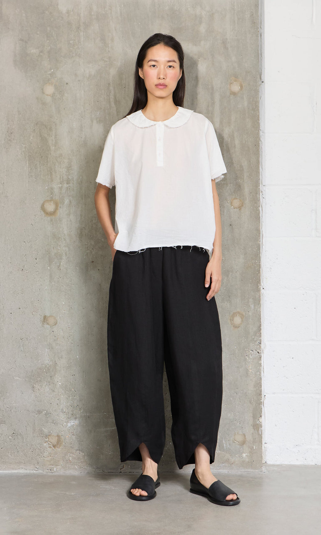 Plümo- A woman in a white blouse and Cazalla Pants - Black stands against a concrete wall, hands in pockets.