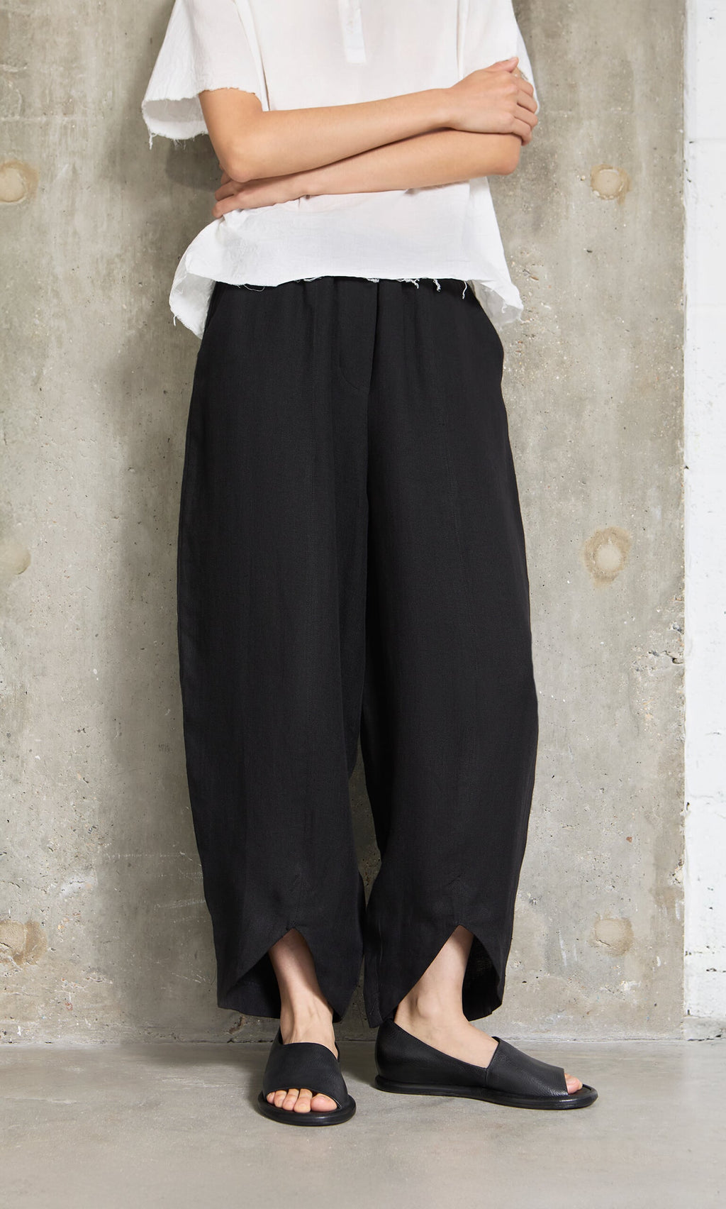 Plümo- Person in a white top and Cazalla Pants - Black stands against a concrete wall with arms crossed.