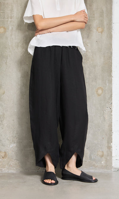 Plümo- Person in a white top and Cazalla Pants - Black stands against a concrete wall with arms crossed.