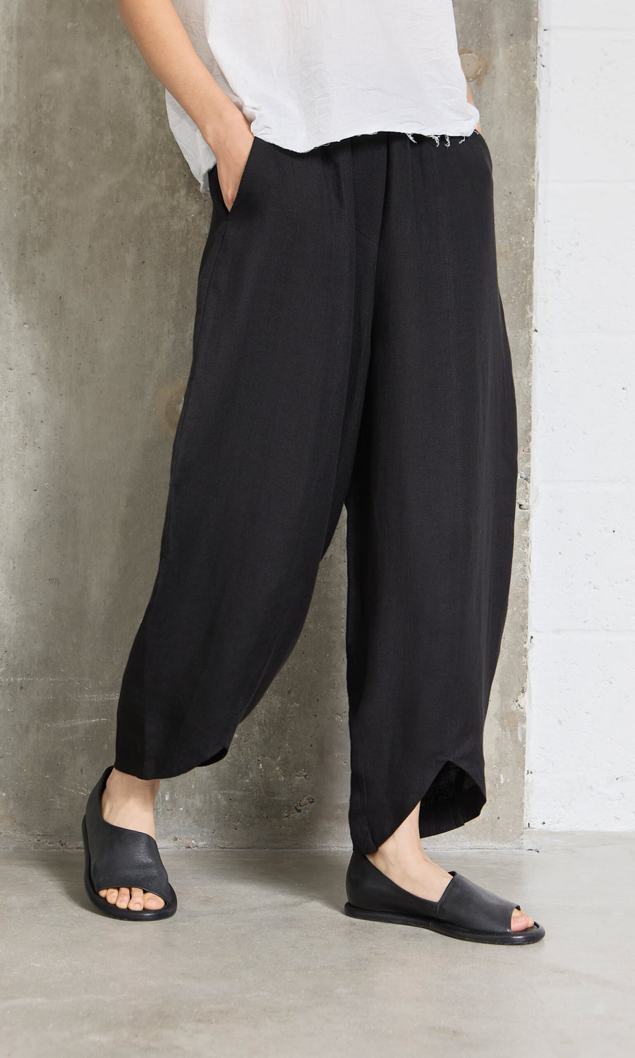 Plümo- Person wearing Cazalla Pants - Black with open-toe sandals, standing against a concrete and white wall.