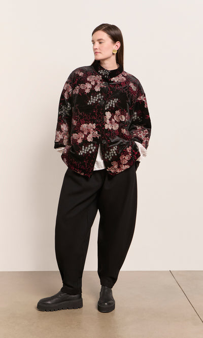 Plümo  Person in loose black pants, black boots, and the Charone Jacket stands against a plain light background.