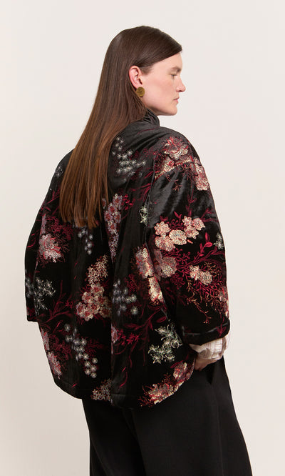 Plümo  Woman in profile wearing the Charone Jacket, a velvet Chinoiserie style with embroidered chrysanthemums, gold earrings.
