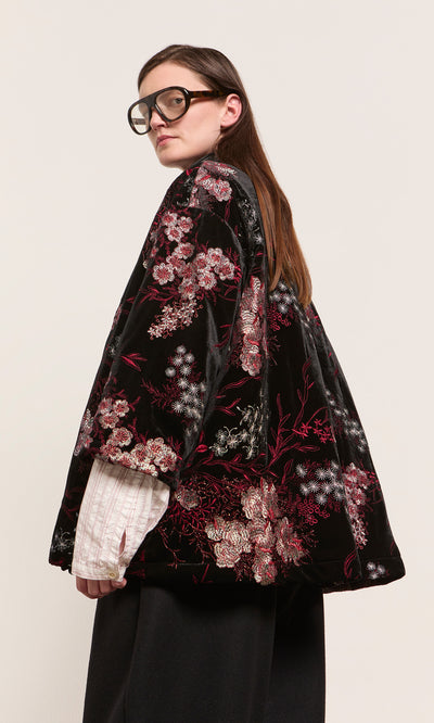 Plümo  Woman in Charone Jacket with embroidered chrysanthemums, white shirt, black pants, round glasses, sideways.