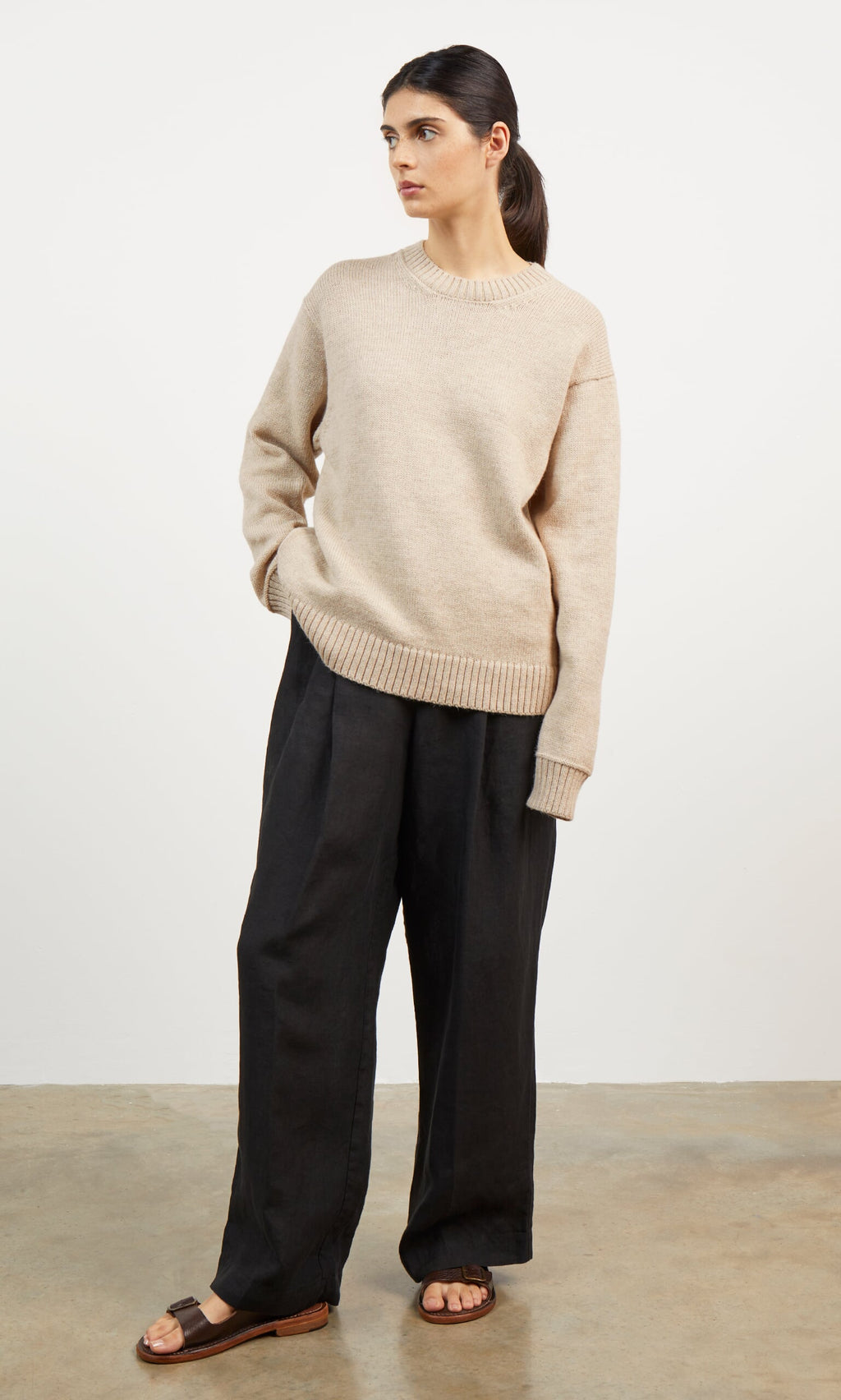 Plümo- Woman in beige Christie Wool Jumper, black wide-leg pants, and brown sandals stands against a white background.