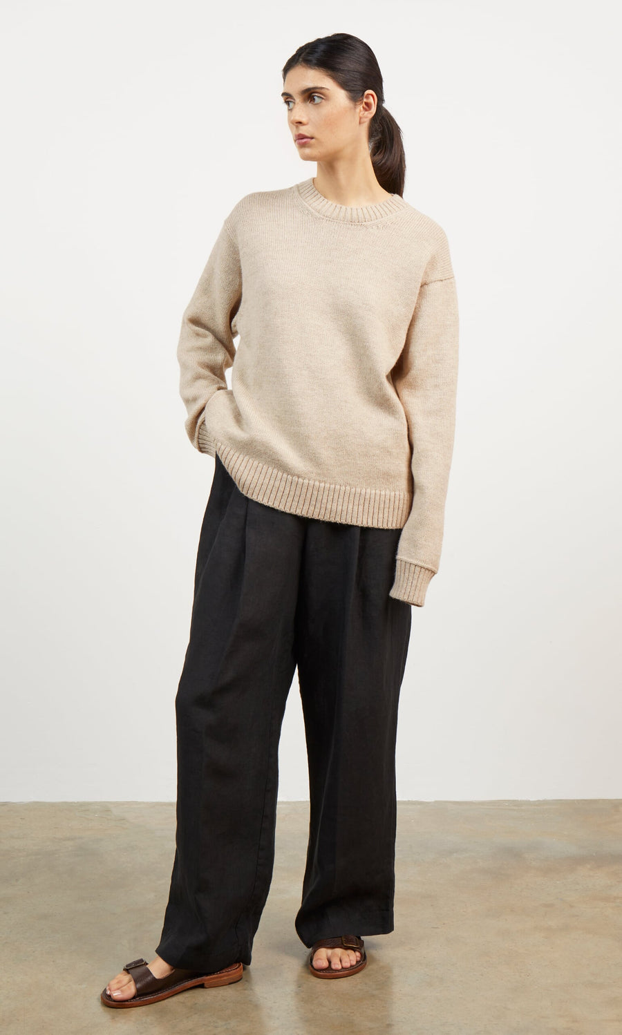 Plümo- Woman in beige Christie Wool Jumper, black wide-leg pants, and brown sandals stands against a white background.