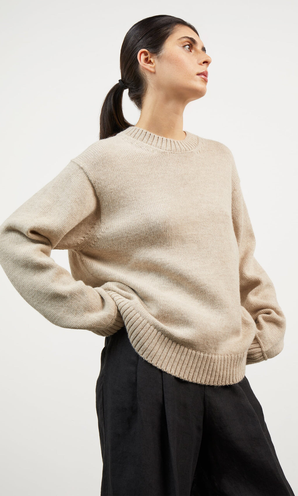 Plümo- Woman wears the Christie Wool Jumper and black pants, standing with hands on hips, looking to the side.