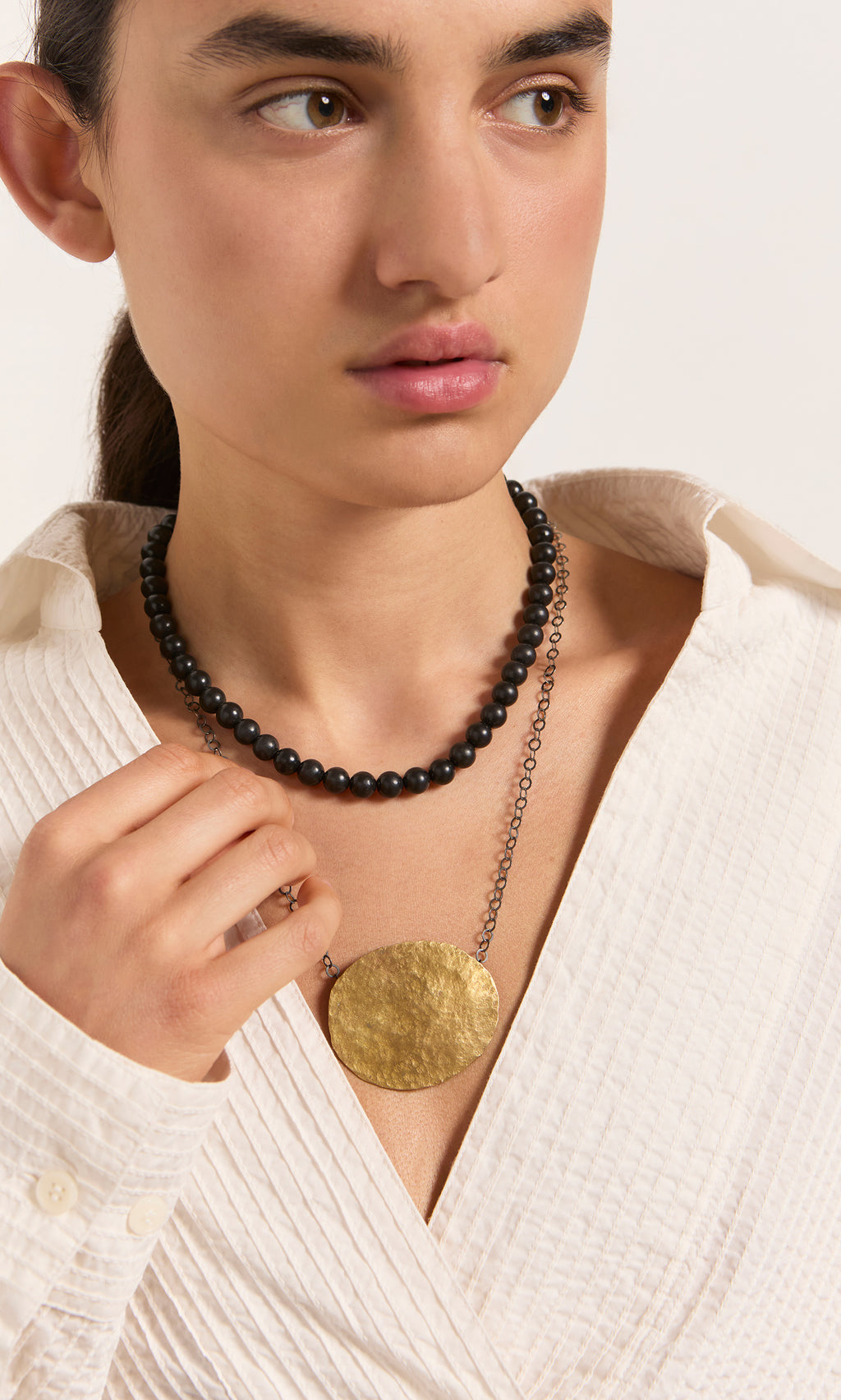 Plümo  Woman in a white textured shirt wears the artisan-made brass Cressa Necklace and gold pendant, looking to the side.