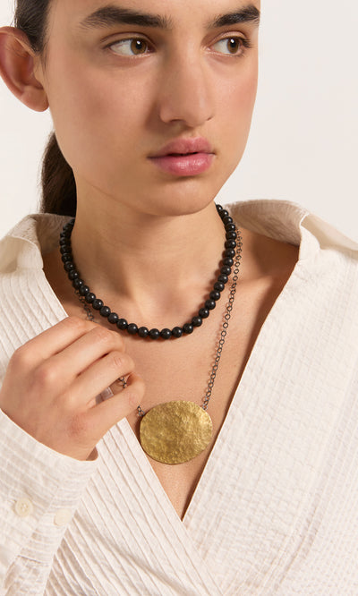 Plümo  Woman in a white textured shirt wears the artisan-made brass Cressa Necklace and gold pendant, looking to the side.