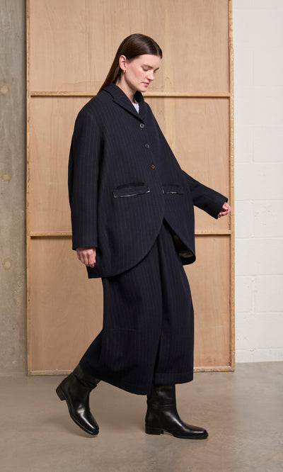 Plümo  A model wears the Cleo Blazer, an oversized navy pinstripe wool style, paired with wide-leg trousers and black boots.