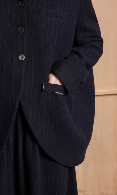 Plümo  A person in the Cleo Blazer with one hand in their pocket stands against a wooden background.