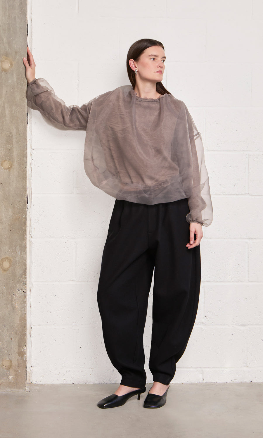 Plümo  Woman in the Clyde Blouse and loose black pants stands against a white brick wall, one hand resting on concrete.