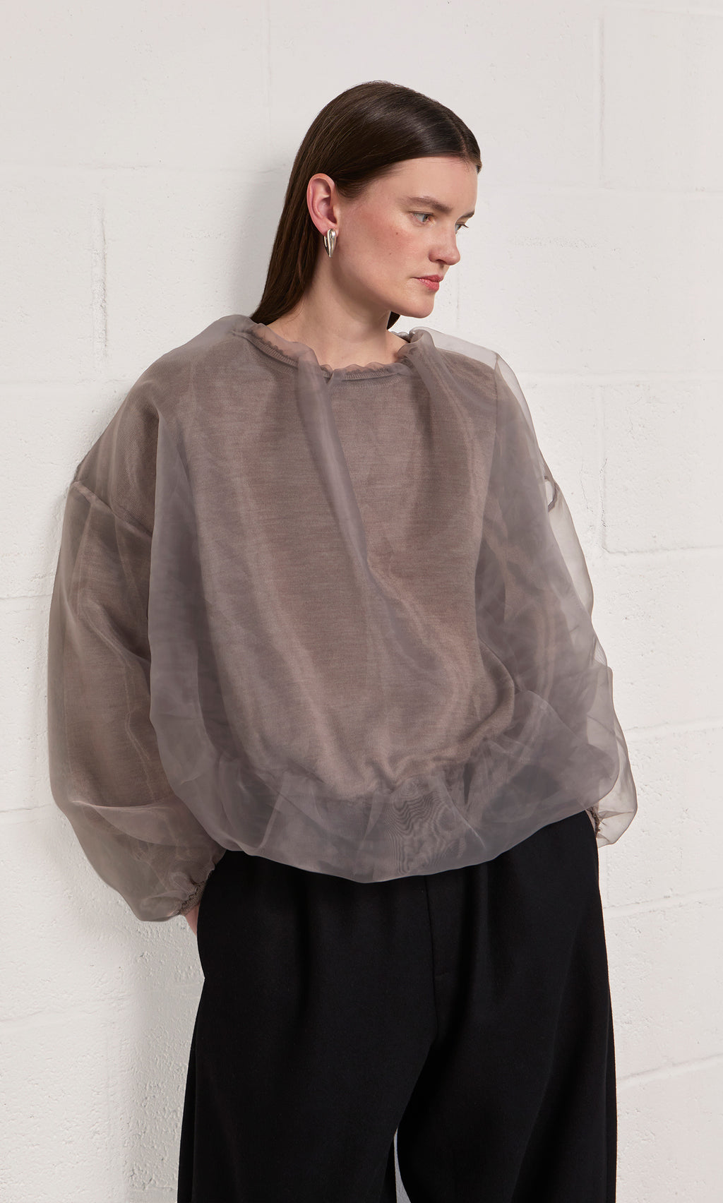 Plümo  Woman wears the Clyde Blouse and black pants, standing by a white textured wall. Made in Italy.