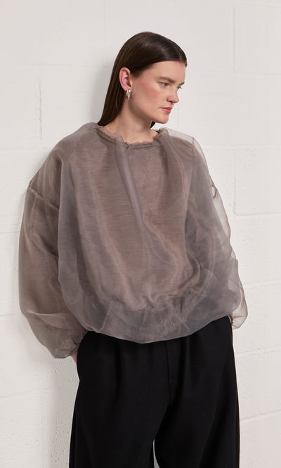 Plümo  Woman wears the Clyde Blouse and black pants, standing by a white textured wall. Made in Italy.