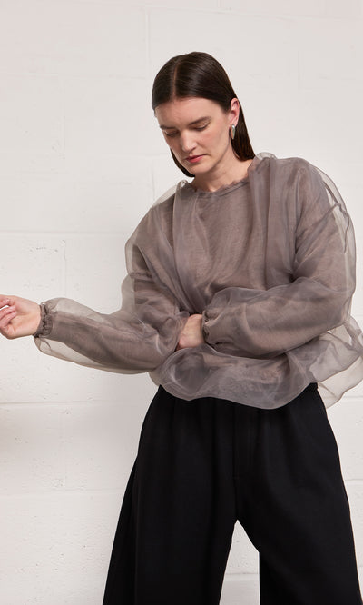Plümo  Woman in the sheer Clyde Blouse and black wide-leg pants stands against a white wall, looking down.