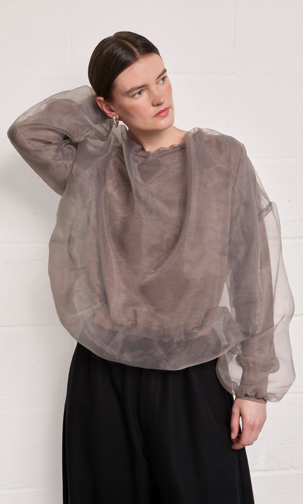 Plümo  Someone in the Clyde Blouse and black pants stands by a white wall, hand resting on their head.