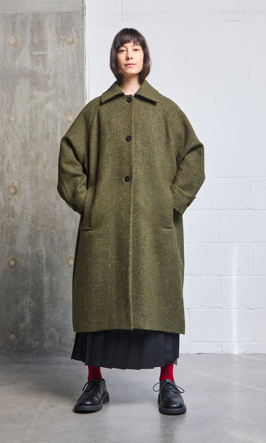 Plümo- A person in an oversized Cormack Coat - Harris Tweed, black skirt, red socks, and black shoes stands by a concrete wall indoors.