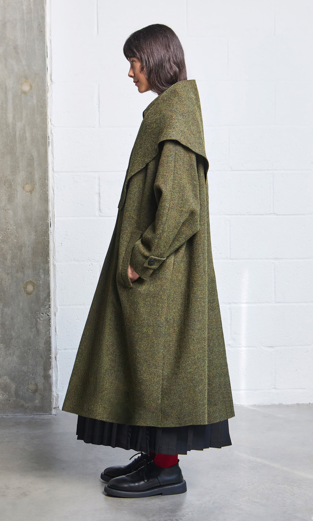 Plümo- Woman in a long Cormack Coat - Harris Tweed, olive-green, wide collar, with black skirt, red socks, black shoes.