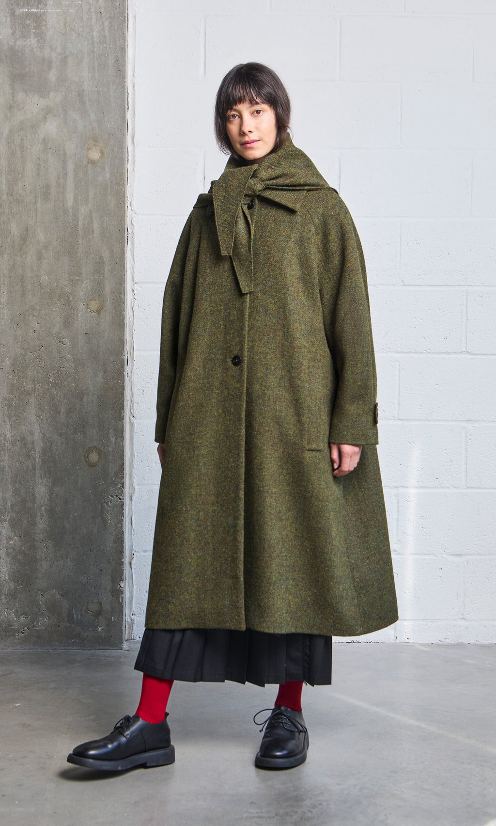 Plümo- A woman wears the Cormack Coat - Harris Tweed in green with a tie collar, standing in a modern industrial space.