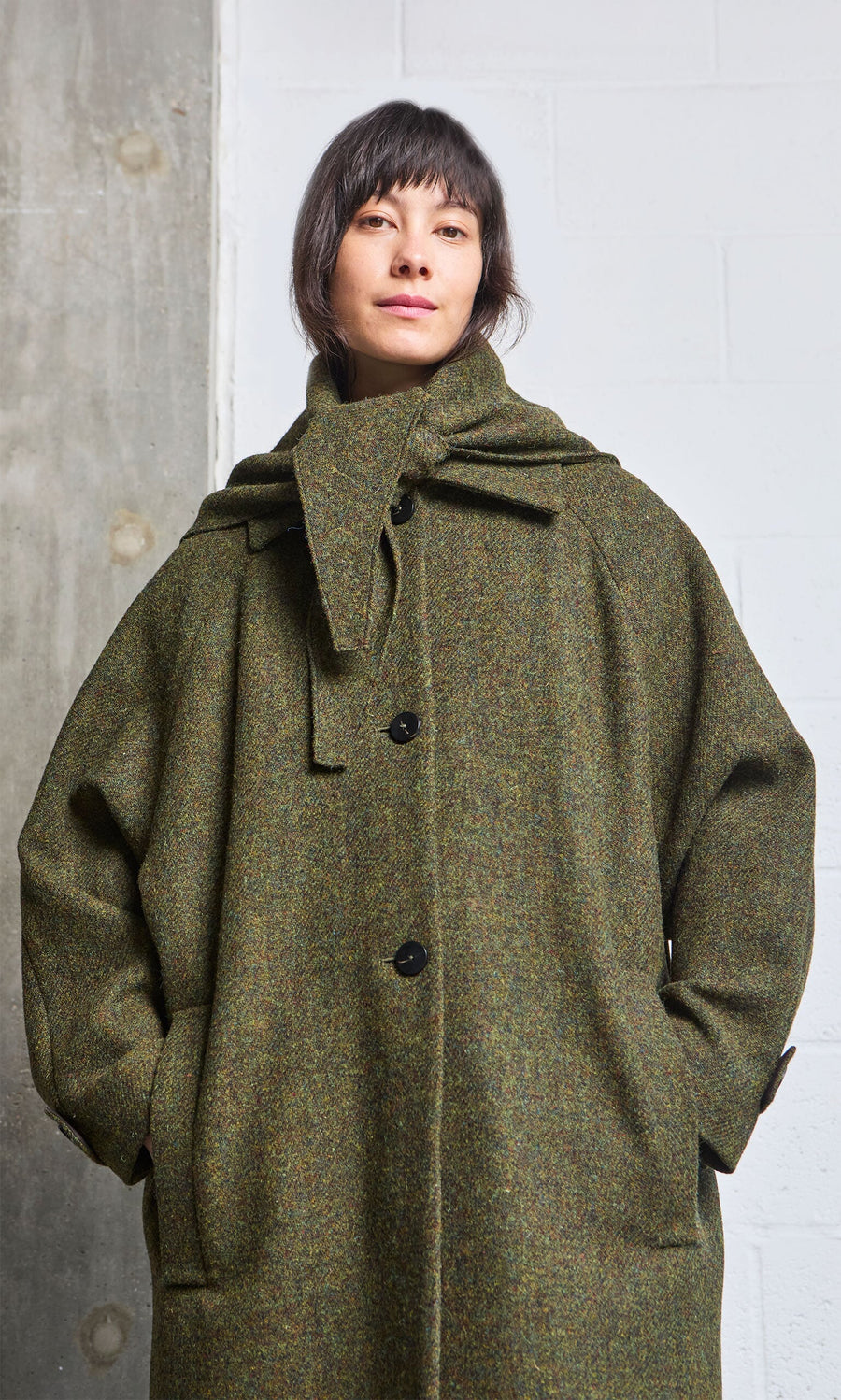 Plümo- Woman wears oversized green Cormack Coat - Harris Tweed with hands in pockets, against a white concrete wall.