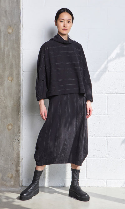 Plümo- A person wears the Speckle Skirt with black boots, standing against a white brick wall, expression neutral.