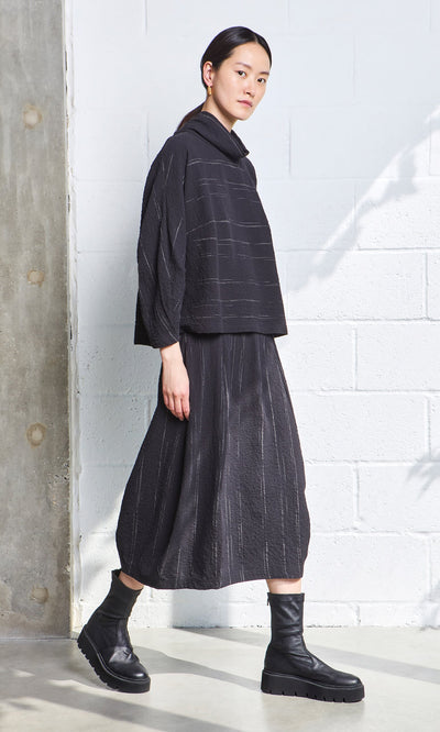 Plümo- Woman in a loose black Cowl Neck Blouse and skirt with black boots, standing by a white textured wall in natural light.