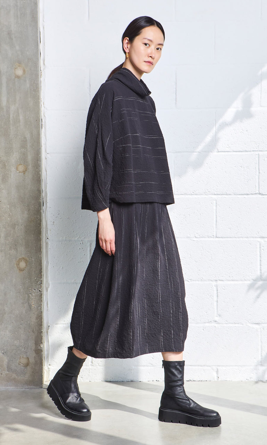 Plümo- Woman in a loose black Cowl Neck Blouse and skirt with black boots, standing by a white textured wall in natural light.