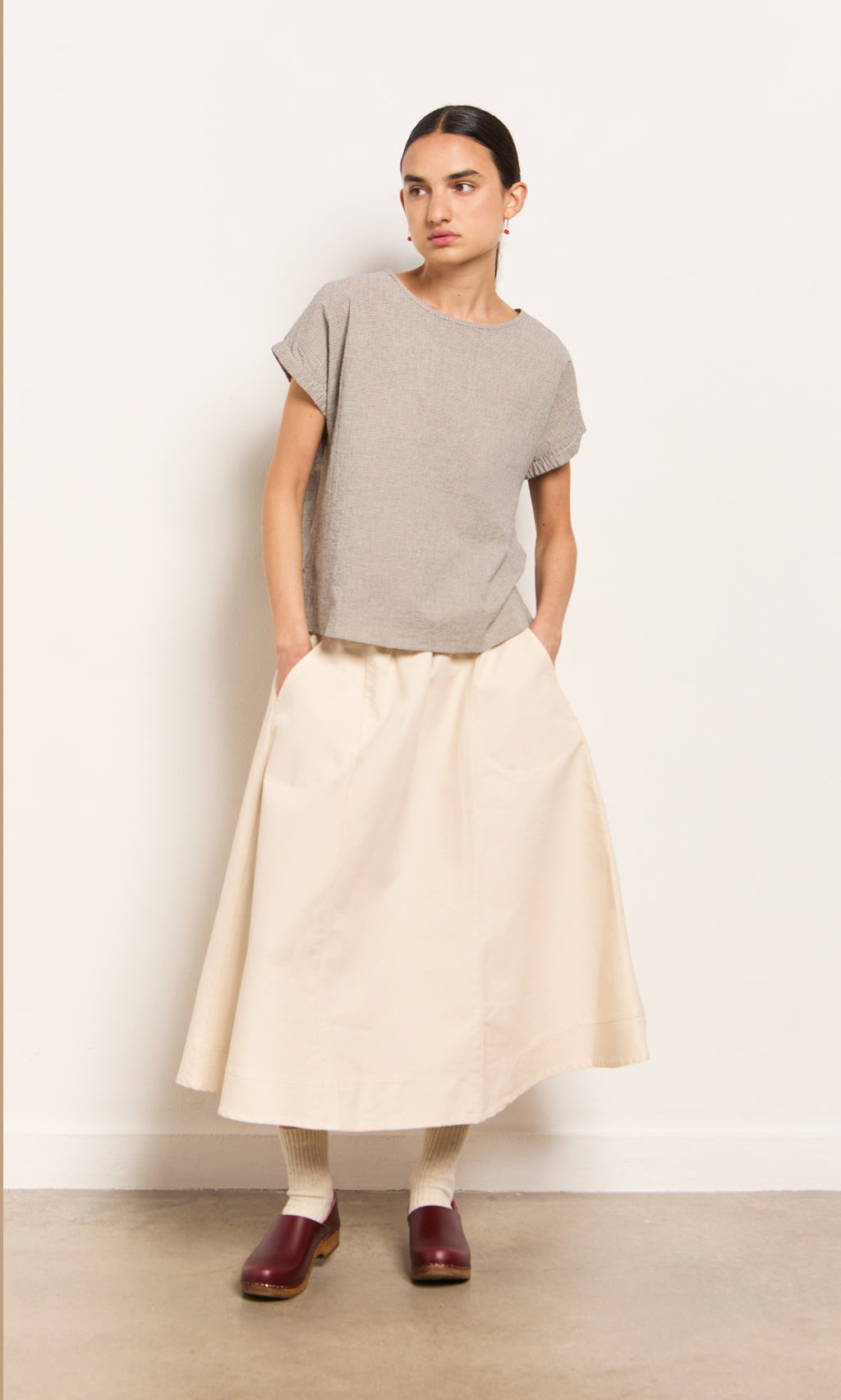 Plümo- Woman in a Crawford Top, beige A-line skirt, red clogs, and socks stands against a plain wall.