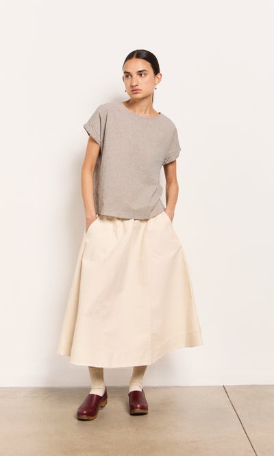 Plümo  Woman in a short-sleeve gray top, Lee Skirt, beige socks, and red clogs stands against a plain white wall.