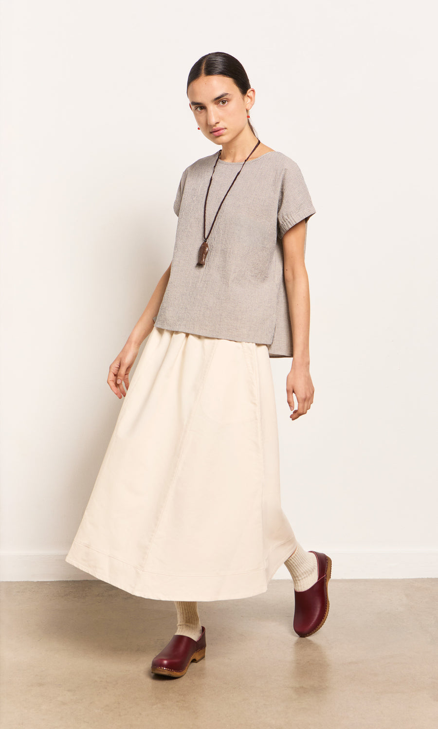 Plümo- Woman wearing the Crawford Top, cream long skirt, maroon clogs, white socks, and a long necklace against a plain background.