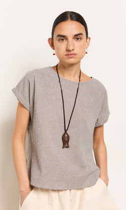 Plümo- Person with dark hair in a low bun wears the Crawford Top, cream pants, and a long fish-shaped pendant necklace.