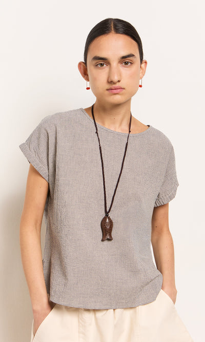 Plümo- Person with dark hair in a low bun wears the Crawford Top, cream pants, and a long fish-shaped pendant necklace.