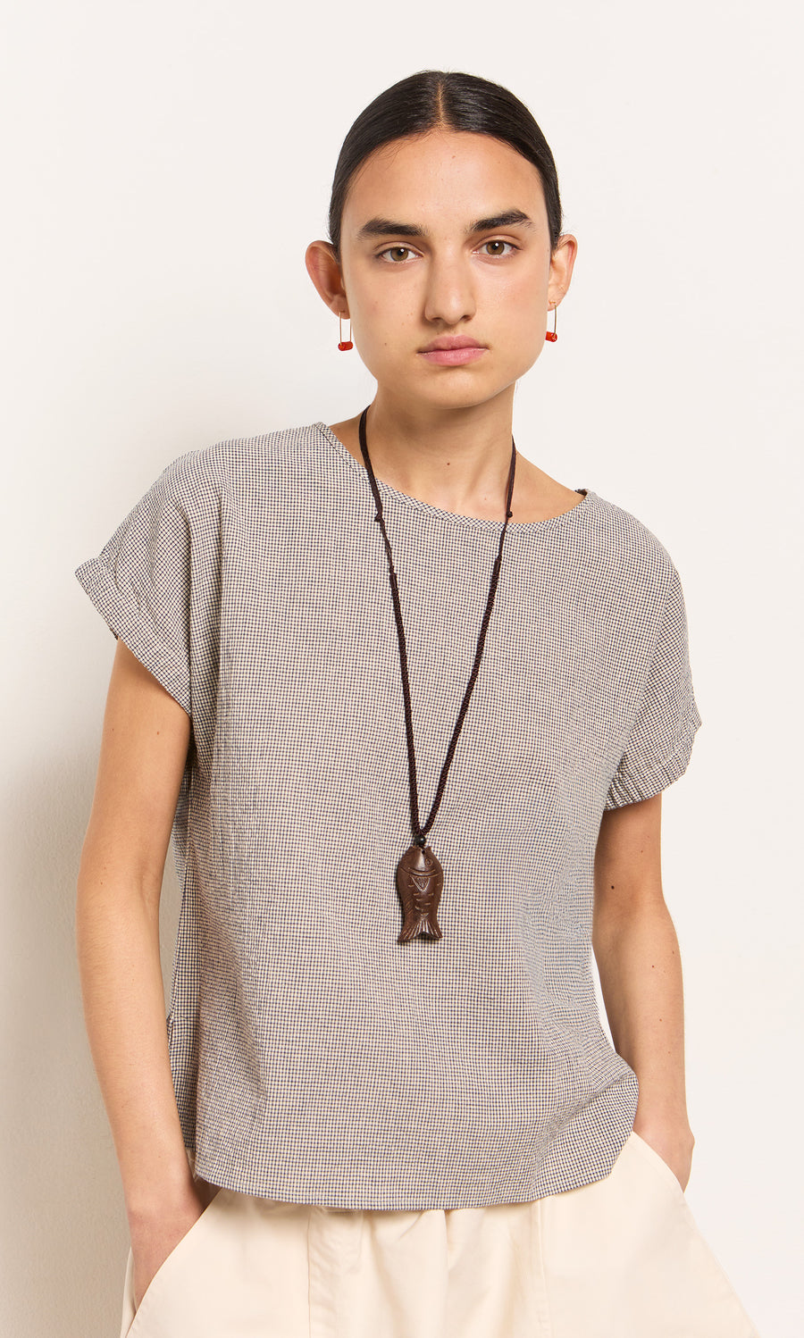 Plümo- Person with dark hair in a low bun wears the Crawford Top, cream pants, and a long fish-shaped pendant necklace.